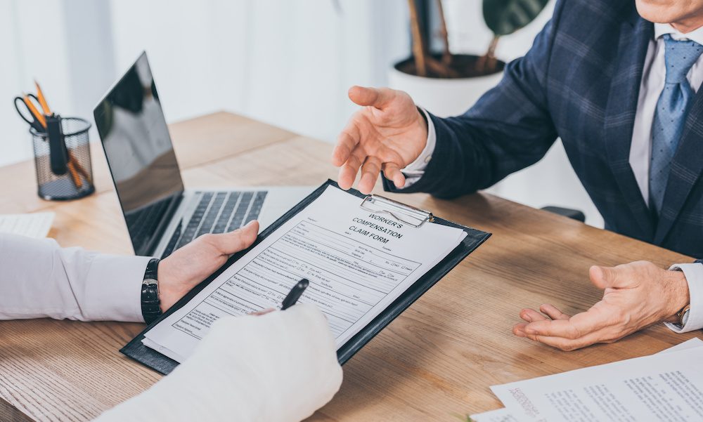 cropped view of worker with broken arm signing form for compensation claim opposite to businessman in blue jacket in office, compensation concept